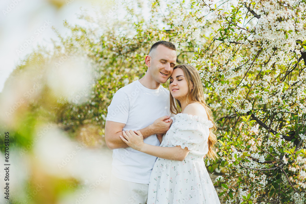 Fototapeta premium A beautiful young couple expecting a pregnancy in a romantic place, a spring blossoming apple orchard. Happy joyful couple enjoying each other while walking in the garden. A man holds a woman's hand
