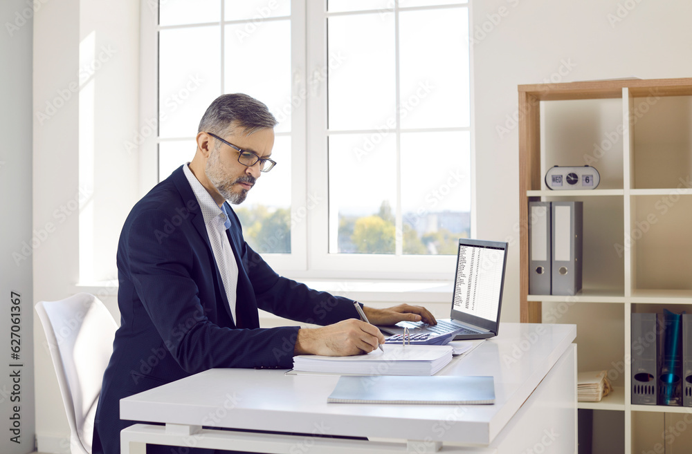 Male accountant or bookkeeper working in office. Mature man in suit and glasses sitting at desk, using modern laptop computer, working with business data, and taking notes in accounting book Male accountant or bookkeeper working in office. Mature man in suit and glasses sitting at desk, using modern laptop computer, working with business data, and taking notes in accounting book