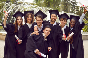 Group photo of happy joyful diverse multiracial college or university graduate student friends in black graduation hats and gowns, with diplomas in hands standing together, with fountain in background