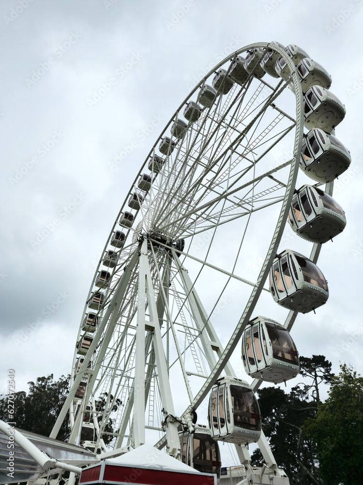 Fototapeta premium photograph of a very tall white Ferris wheel, with fifty cabins. in the background the sky with many clouds, and the foliage of some trees in a park