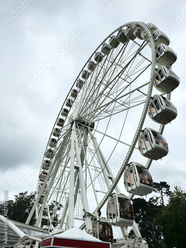 photograph of a very tall white Ferris wheel, with fifty cabins. in the background the sky with many clouds, and the foliage of some trees in a park