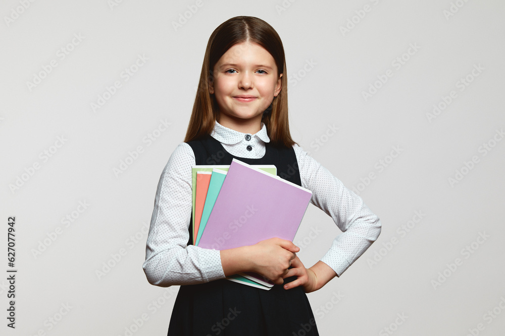 Adorable positive little girl in school uniform holding textbooks while looking at camera against white background. Back to school concept Adorable positive little girl in school uniform holding textbooks while looking at camera against white background. Back to school concept