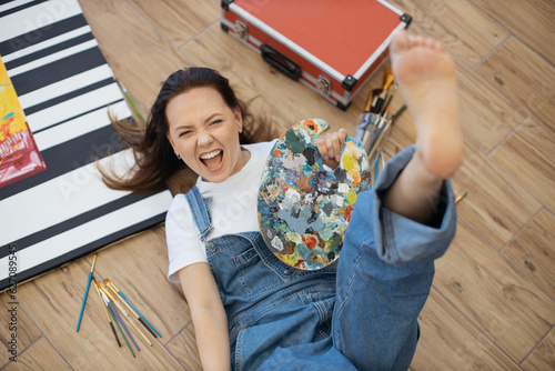 Bilde på lerret Excited female artist in denim outfit screaming cheerfully with raised leg on wooden floor of modern studio