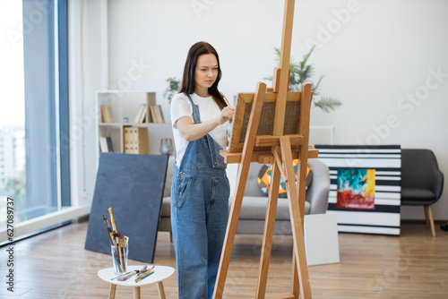 Selective focus of beautiful lady in oversized jumpsuit holding brush and palette while staying in studio. Charming young woman defining new elements on canvas with oil paints using direct lighting.