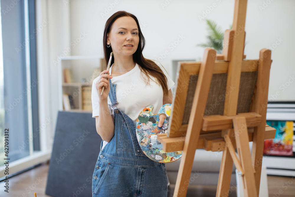 Portrait of talented brunette woman posing with paintbrushes on cheek and color mixing palette in hand. Inspired creative in white T-shirt and jeans using daylight in studio space for productive work. Portrait of talented brunette woman posing with paintbrushes on cheek and color mixing palette in hand. Inspired creative in white T-shirt and jeans using daylight in studio space for productive work.