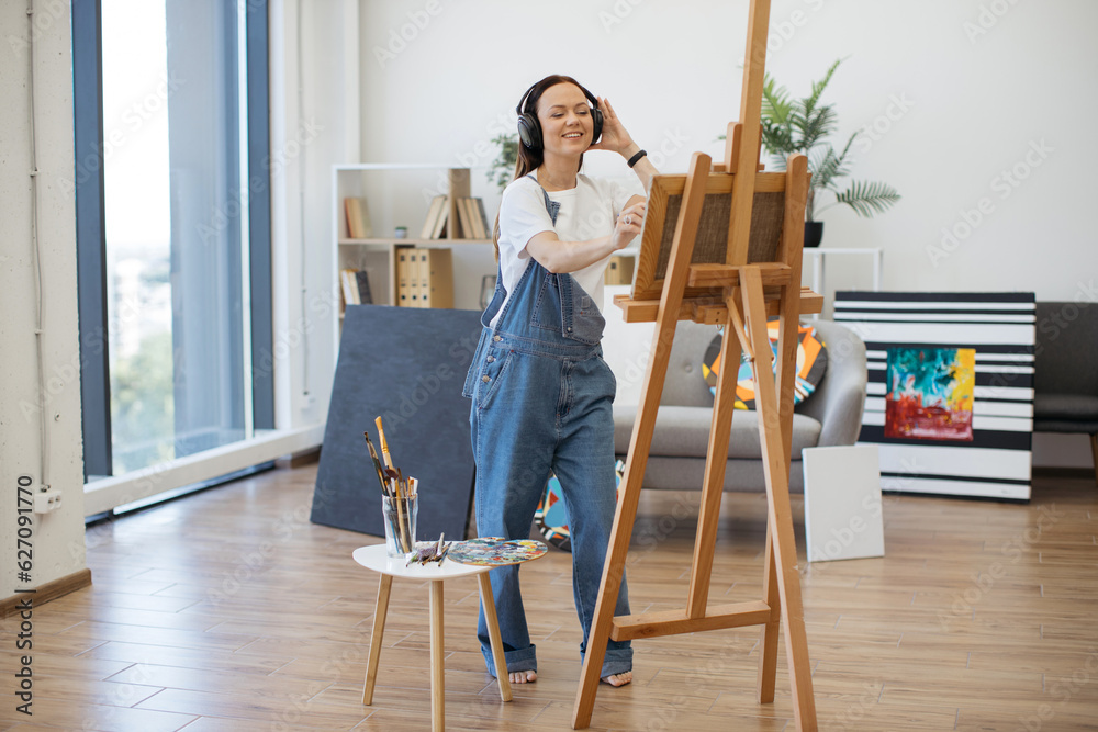 Happy brunette artist in denim jumpsuit handling brush professionally while rocking out to tunes in hands-free device. Easygoing woman approaching visual design under influence of melodies indoors. Happy brunette artist in denim jumpsuit handling brush professionally while rocking out to tunes in hands-free device. Easygoing woman approaching visual design under influence of melodies indoors.