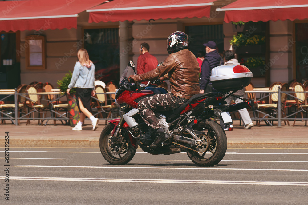 A motorcyclist in a helmet rides through the city on a red sports touring motorcycle. A man travels on a motorbike through a beautiful city next to the sidewalk.