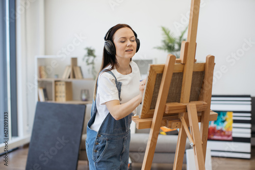 Positive young lady in denim jumpsuit moving to beat of song in headphones with wide brush in hand. Fine-artist enriching imagination with emotions while taking pleasure of sound quality via device.