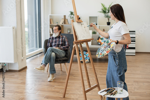 Side view of caucasian lady in cozy clothes applying paint on cotton canvas while keeping eyes on sitter in studio space. Talented amateur artist getting experience by drawing from calm senior man.