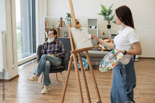 Relaxed older man posing in comfortable chair while young woman with palette applying paint on canvas. Avid arts student making initial drawing during portrait painting lesson in studio space.