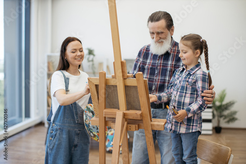 Caring older adult embracing granddaughter near artist easel while young woman fixing paper on canvas. Cute talented girl learning about new textures and shapes during art lesson with mom at home.