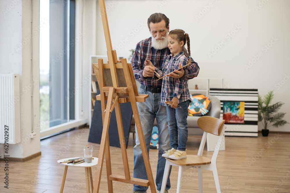Full length view of grey-haired man preparing paint on palette while stylish girl standing in his hug. Relaxed child in denim outfit taking pleasure of art making with grandpa in studio at home.