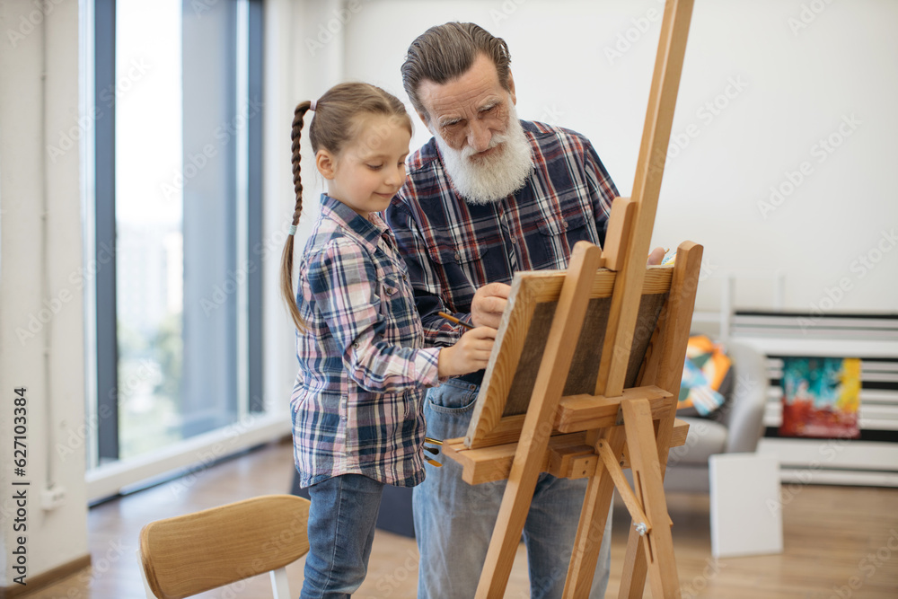 Grey-haired man preparing paint on palette while stylish girl standing in his hug. Relaxed child in denim outfit taking pleasure of art making with grandpa in studio at home.