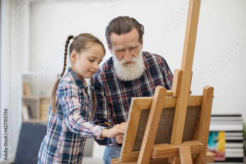 Close up view of senior grandfather and little child in similar outfits working on interesting art project. Caring grandpa teaching how to fill drawing in with color from palette in home studio.