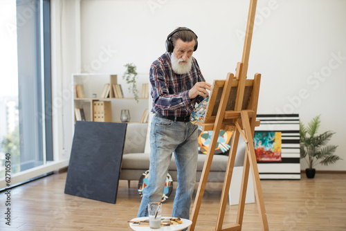 Full length view of aged gentleman wearing headphones making brush strokes while standing near easel indoors. Experienced artist drawing inspiration for work from relaxing music in studio space.