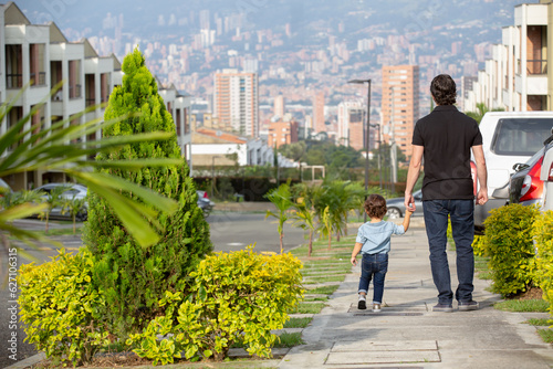 Back of a father and small daughter holding hands while walking on a sidewalk around their home in a residential area.