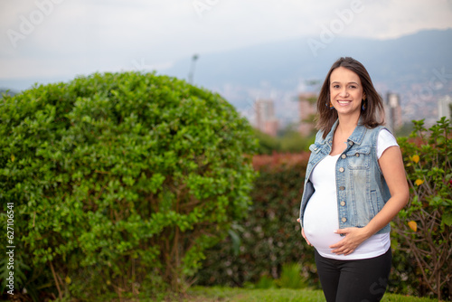 Happily smiling pregnant mother in a nature landscape with some city buildings and mountains, hands holding her belly.