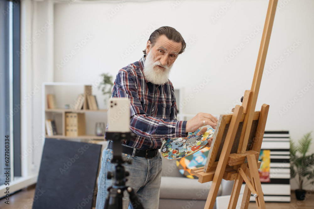 Caucasian creative painter standing with drawing palette next to wooden easel and working on new portrait. Mature man making video recording on white modern smartphone using tripod. Caucasian creative painter standing with drawing palette next to wooden easel and working on new portrait. Mature man making video recording on white modern smartphone using tripod.