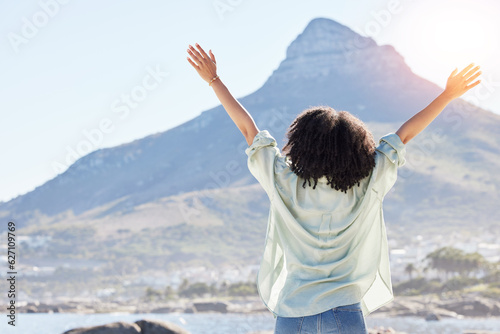 Woman at beach, arms in air and freedom, back view with mountains and travel, praise and sunshine. Female person outdoor, carefree and adventure, mindfulness and peace with worship in nature