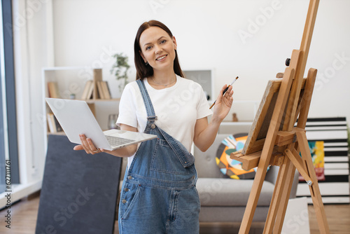 Portrait of friendly expert in art posing on camera with personal laptop and brush and frankly smiling at home. Positive beautiful woman in casual outfit drawing picture from internet on canvas.