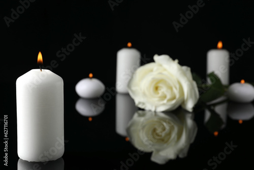 White rose and burning candles on black mirror surface in darkness  closeup with space for text. Funeral symbols