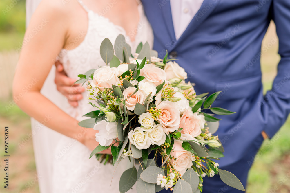 A bride and groom are standing together with their arms around each other, and she holds a soft pink and white classic wedding bouquet, and he wears a blue suit jacket. A bride and groom are standing together with their arms around each other, and she holds a soft pink and white classic wedding bouquet, and he wears a blue suit jacket.