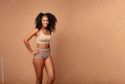 A beautiful ebony model in a sporty slim top and shorts stands on a beige background with copy space.