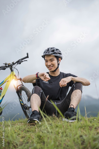 happy young Asian man in sportswear checks his calories burned on his smartwatch while resting on the grass after a long bike ride in the mountains.