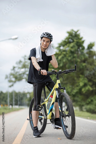 A tired and sweaty young Asian man in sportswear and a bike helmet wiping his neck with a towel while biking along the country roads.