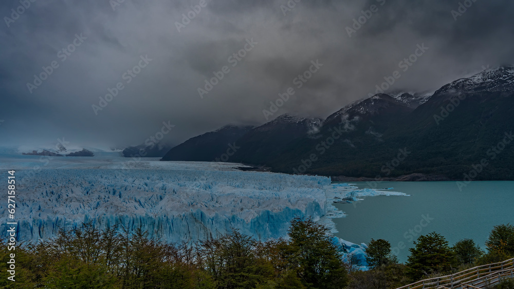 From the observation deck you can see the endless blue glacier Perito Moreno and turquoise lake. Green vegetation in the foreground. Mountains in fog and clouds. Argentina. El Calafate.