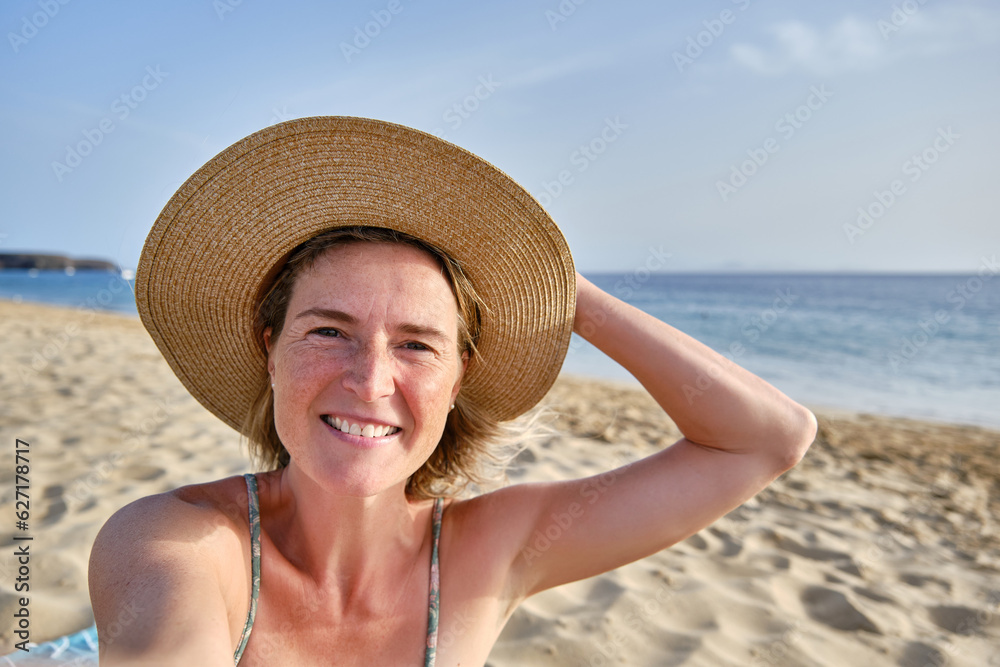 Smiling woman in hat standing on sandy beach and taking selfie