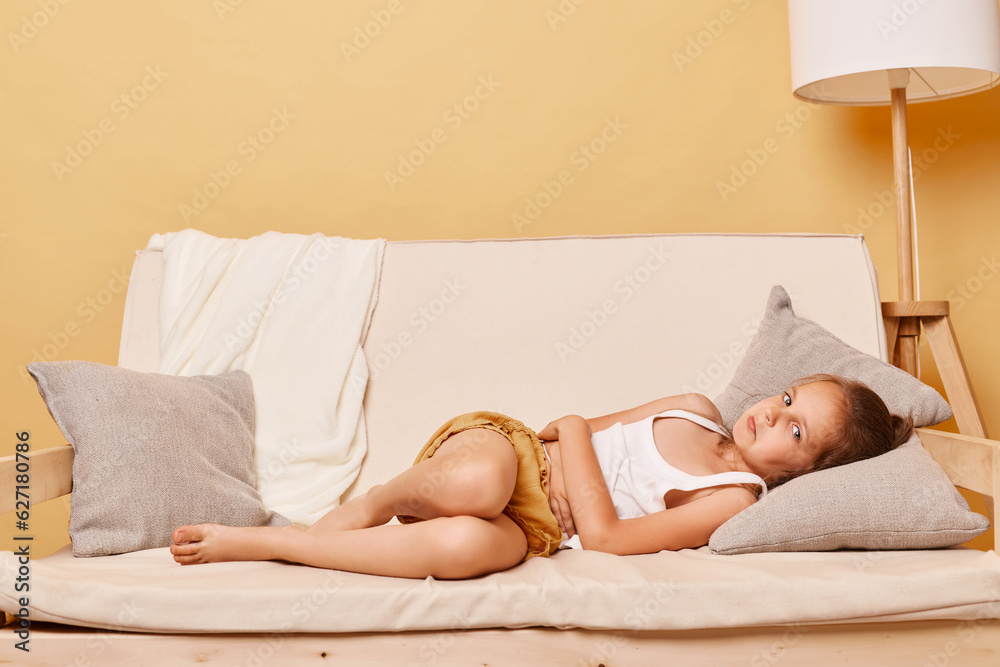 Obraz premium Sad sick little girl wearing casual clothing sitting on sofa at home against beige wall lying on couh touching her painful stomachache looking at camera with pout lips.