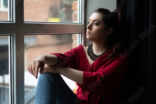 a beautiful young girl in a red shirt with a handmade jewelry necklace around her neck is posing near the window