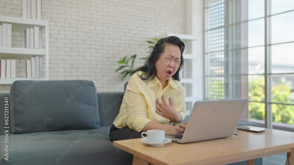 Elderly asian woman having chest pain while working on laptop at home. Loving daughter take care of elderly mother while having left chest pain on sofa. Suffering from chest pain or heart attack