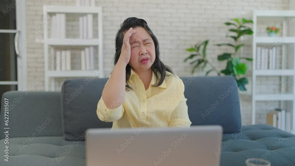 Stressed elderly asian woman having headache while working on laptop at home. Daughter giving medicine to mother having a headache pain. Elderly health problems