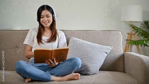 Young Asian woman with headphones sitting on the sofa, relaxing while reading a book