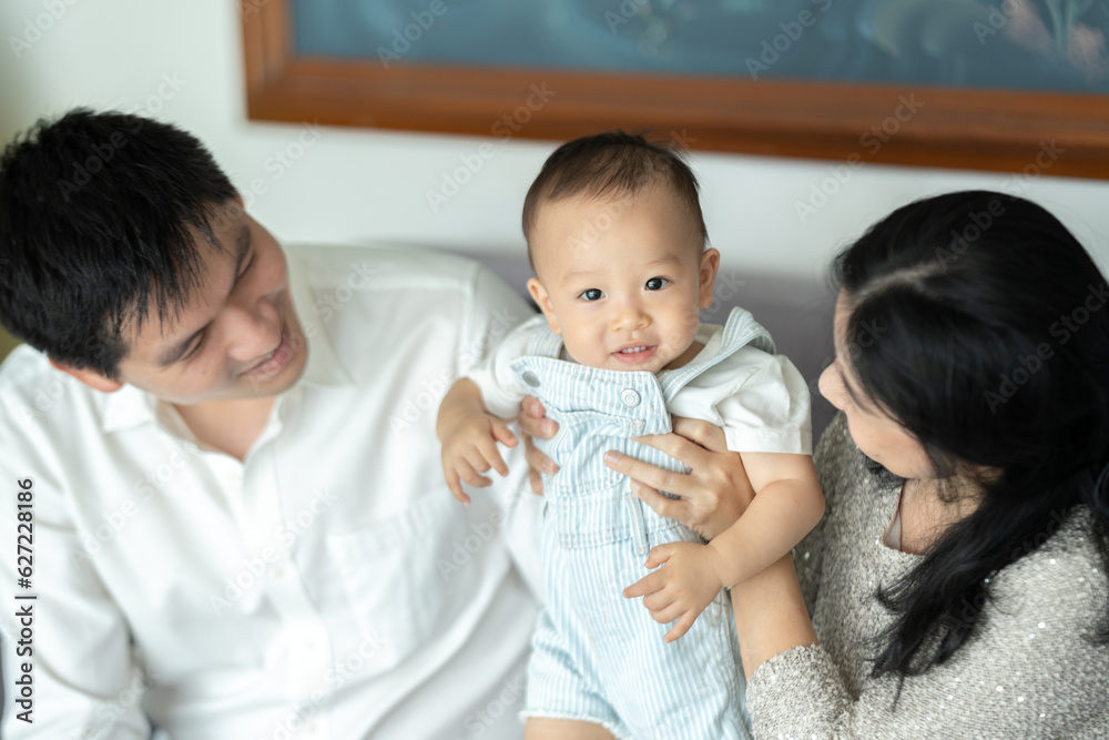 Young and cheerful mother and father sitting on couch relaxing while playing with cute little new born baby boy and toddler who is trying to stand with support at home during weekend Young and cheerful mother and father sitting on couch relaxing while playing with cute little new born baby boy and toddler who is trying to stand with support at home during weekend