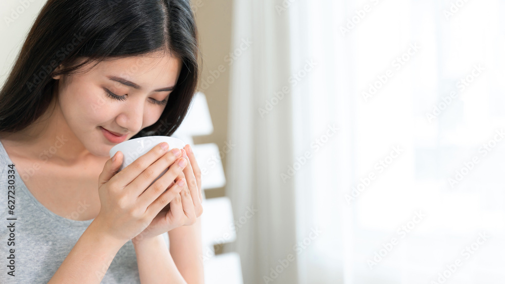 Asian beautiful woman sitting on bed in the bedroom and holding coffee cup in hand with happy Asian beautiful woman sitting on bed in the bedroom and holding coffee cup in hand with happy