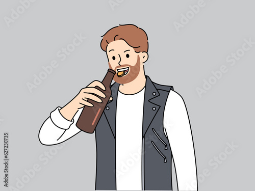 Brutal man opens bottle of beer with teeth and smiles, suffering from alcohol addiction. Young bearded guy in leather vest drinks beer enjoying taste of refreshing intoxicating drink