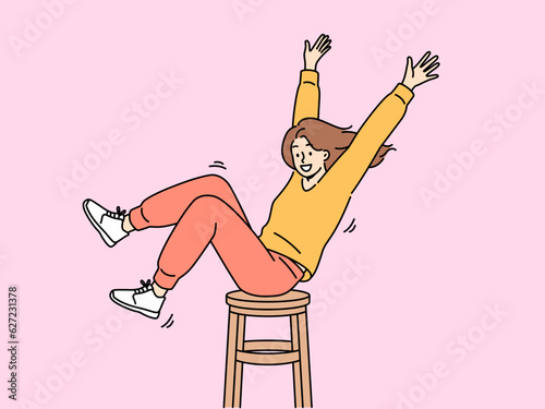 Happy girl is spinning on wooden chair, raising hands up and sharing good mood with others. Positive teenager girl in stylish clothes enjoys life without wanting to pay attention to troubles
