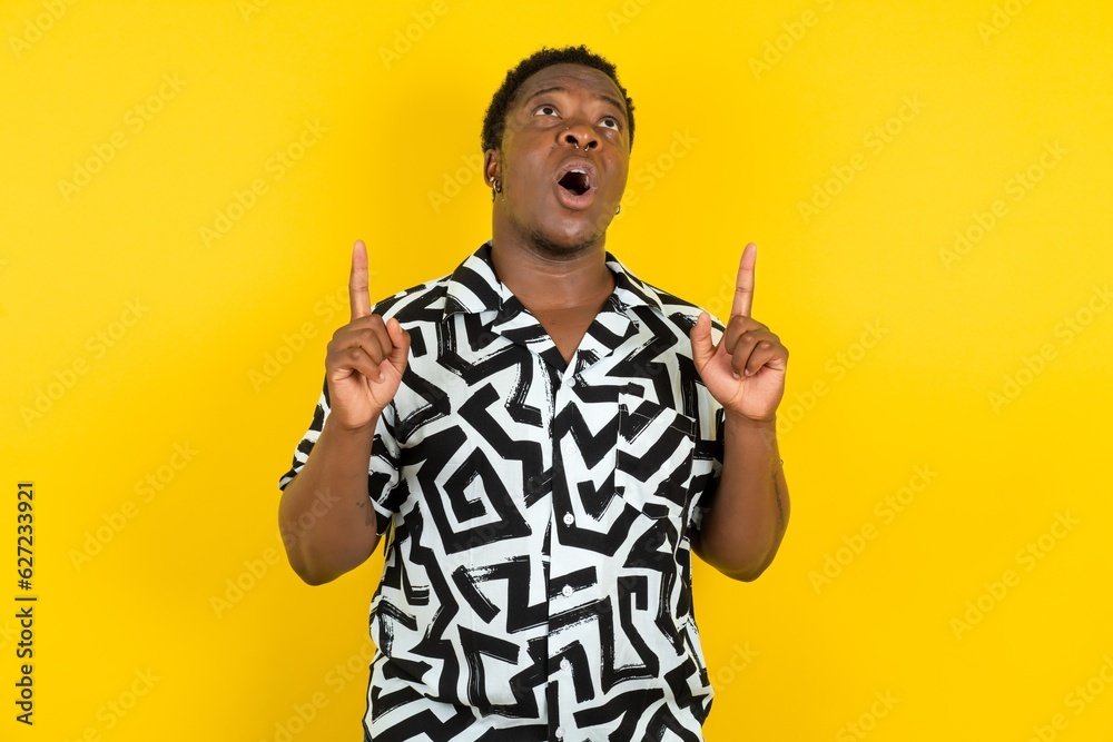 Young latin man wearing printed shirt over yellow background amazed and surprised looking up and pointing with fingers and raised arms.