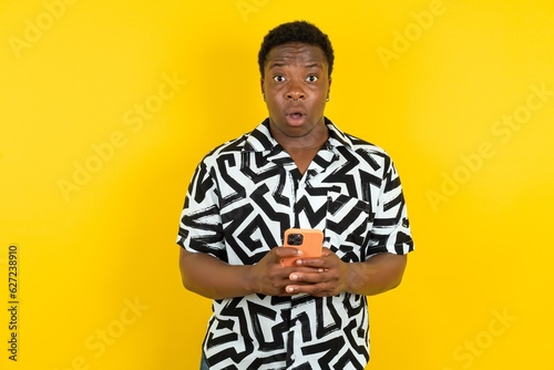 Shocked Young latin man wearing printed shirt over yellow background opens mouth hold phone reading advert unbelievable big shopping prices