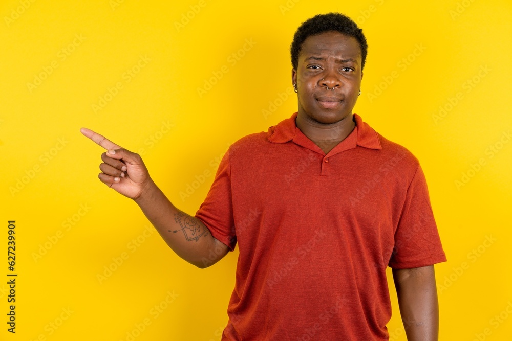 Serious Young latin man wearing red t-shirt over yellow background smirks face points away on copy space shows something unpleasant. Look at this advertisement. Big price concept Serious Young latin man wearing red t-shirt over yellow background smirks face points away on copy space shows something unpleasant. Look at this advertisement. Big price concept