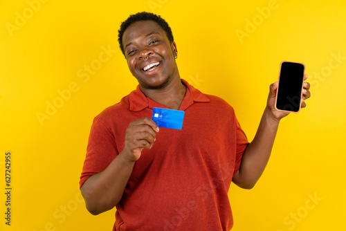 Young latin man wearing red t-shirt over yellow background opened bank account, holding smartphone and credit card, smiling, recommend use online shopping application