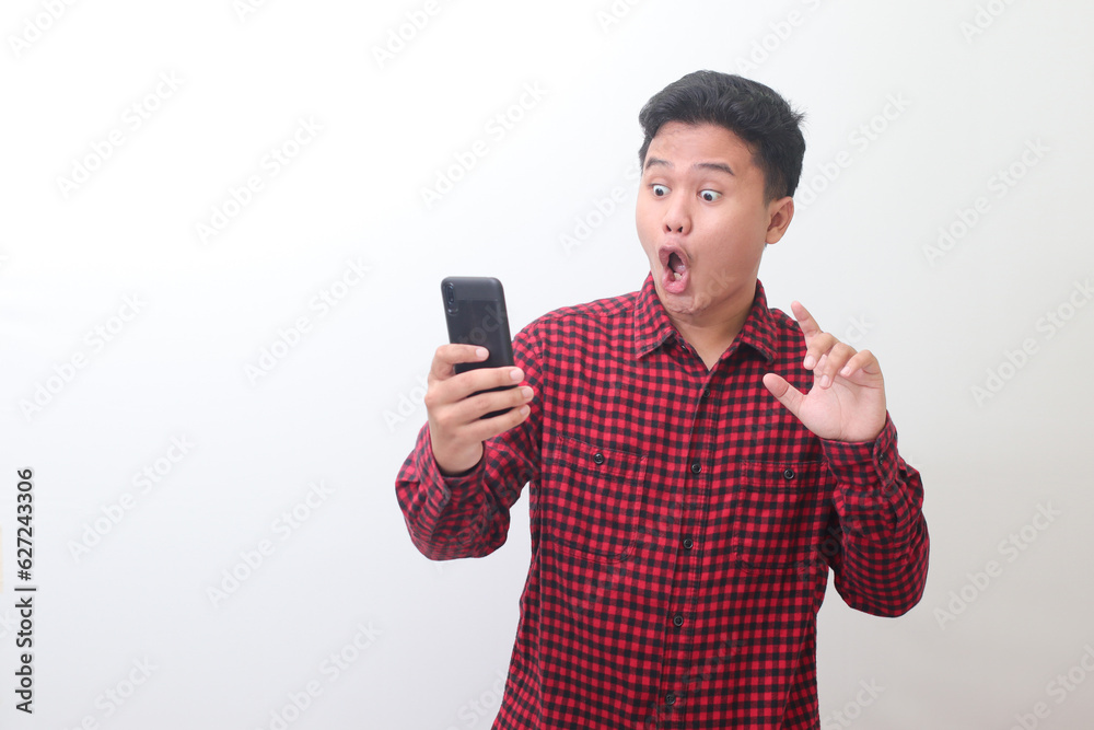 Portrait of shocked Asian man in casual plaid shirt standing against gray background, holding a mobile phone while trying to touch it. Advertising concept. Portrait of shocked Asian man in casual plaid shirt standing against gray background, holding a mobile phone while trying to touch it. Advertising concept.
