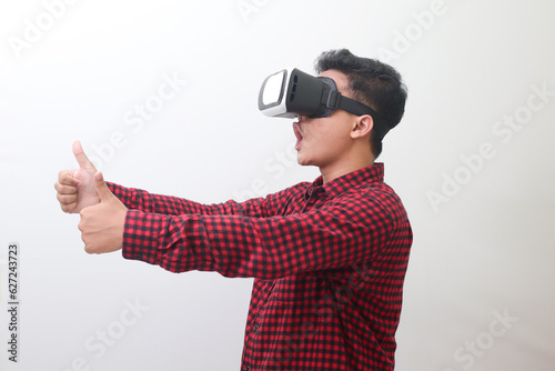 Portrait of Asian man in red plaid shirt using Virtual Reality (VR) glasses and showing thumb up hand gesture. Isolated image with copy space on white background.