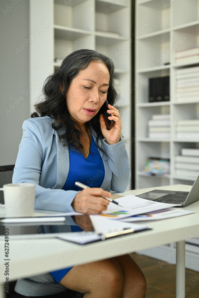Beautiful mature businesswoman having pleased phone conversation and taking notes Beautiful mature businesswoman having pleased phone conversation and taking notes