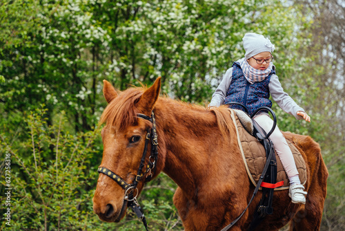 Little girl riding on a horseback. The girl is sitting on a horse . A child on a day walk on the ranch. © Iryna