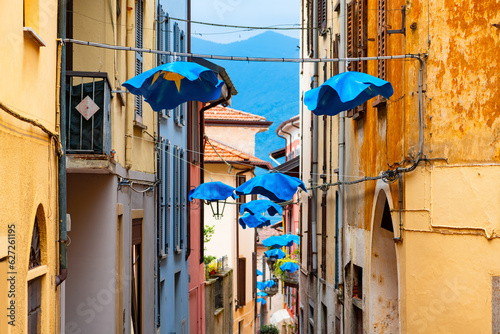 Narrow street in the old town of Luino at lake “Lago Maggiore“ in Italy. Colorful facades, balconys and shops. Popular tourist destination and famous market place on a summer day. Idyllic alley lane.