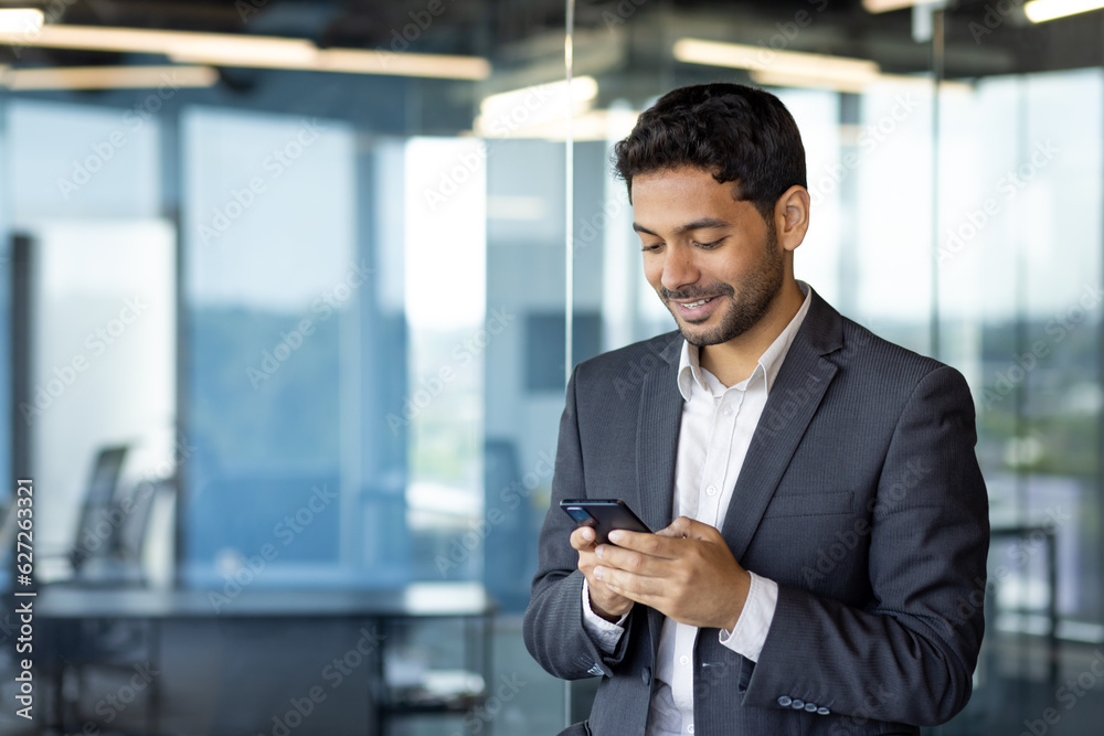 Young arab businessman holding phone, man inside office at workplace typing message and browsing online sites, worker using app on smartphone. Young arab businessman holding phone, man inside office at workplace typing message and browsing online sites, worker using app on smartphone.